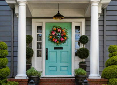 A turquoise coloured front door decorated with a large floral wreath on a traditional home with brick path and trimmed hedges.